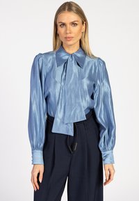 Lichtblauw satijnen blouse met een grote kraag en strikdetail, voorzien van puffed lange mouwen en manchetten, gedragen met donkere broeken.