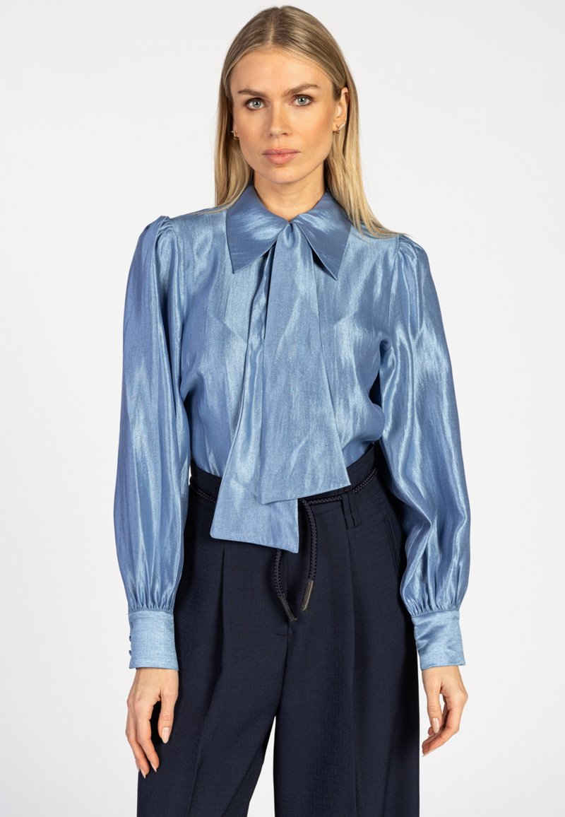 Lichtblauw satijnen blouse met een grote kraag en strikdetail, voorzien van puffed lange mouwen en manchetten, gedragen met donkere broeken.