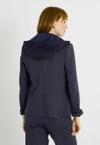 Chaqueta azul marino con patrón y capucha, con un ajuste entallado, tela texturizada y puños remangados para un estilo ajustable.