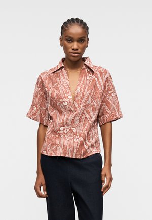 Femme aux cheveux tressés portant un chemisier à manches courtes couleur rouille avec un motif floral blanc et un pantalon sombre sur un fond uni.