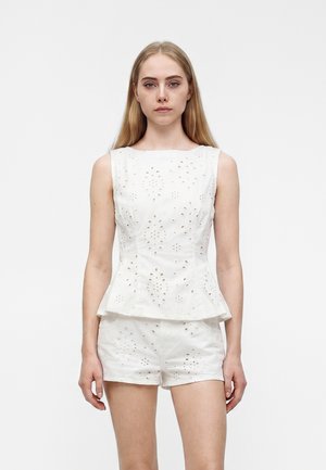 Giovane donna con lunghi capelli biondi che indossa un top bianco senza maniche in tessuto traforato e pantaloncini bianchi traforati abbinati, in piedi davanti a uno sfondo bianco uniforme.