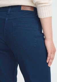Džíny v námořnické modré barvě z denimu se koženým štítkem s nápisem "OXMO" na zadní straně, boční kapsou a přiléhavým střihem. Povrch se jeví jako hladký.