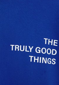 Blauw katoenen stof met een gladde textuur toont witte tekst met de woorden "THE TRULY GOOD THINGS" in verschillende lettergroottes en orientaties.