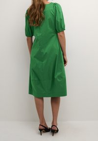 Grön klänning till knäna i lätt tyg, med utsvängda korta ärmar och en enkel silhuett. Bärs med svarta klacksandaler.