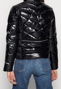 Czarna kurtka puchowa z błyszczącym, wytłaczanym wzorem w zygzaki. Posiada krótki krój oraz oznaczenie marki na pasku z tyłu. W połączeniu z niebieskimi dżinsami.