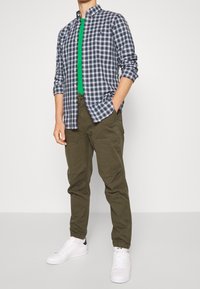 Camisa a cuadros azul y blanca, camiseta verde debajo, pantalones jogger caqui y zapatillas blancas con detalles negros; look casual.