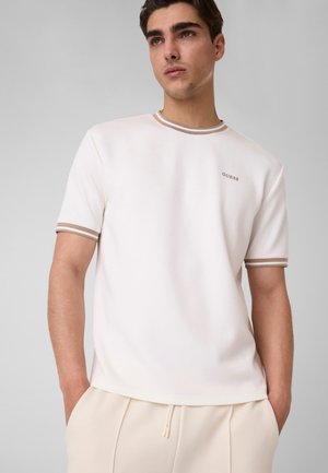 Jeune homme aux cheveux courts et foncés portant une chemise blanche à manches courtes avec une bordure beige et un pantalon beige, les mains dans les poches, regardant ailleurs.