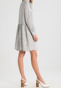 Grå randig klänning med långa ärmar, knappmanschetter och knyt i halsen. Har en utsvängd kjol med en rynkad midja. Kombinerad med vita skor med klack.