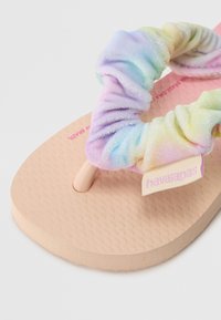 Par de chinelos com tiras de tecido tie-dye em tons pastel, sola de borracha texturizada bege e uma etiqueta de logo rosa com a inscrição "havaianas".