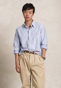 Camisa azul clara com riscas, de botões, com mangas arregaçadas, calças pregueadas em tom bege e um cinto castanho. Relógio de pulso notável visível no pulso esquerdo.