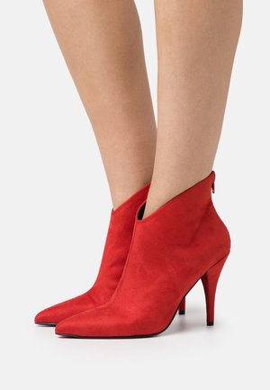 Bottes cheville en daim rouge à bout pointu et talons aiguilles portées sur des jambes nues sur un fond uni.