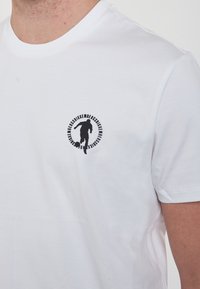 T-shirt in cotone bianco con un logo circolare nero che presenta una figura sportiva e del testo, colletto rotondo, maniche corte e texture morbida.