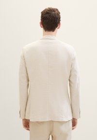 Blazer en lin beige clair avec un design structuré, fente simple à l'arrière et poignets boutonnés. Texture lisse et style minimaliste.