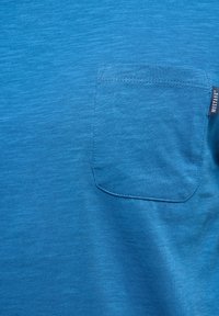 Tissu bleu avec une poche poitrine cousue sur le côté droit, comportant une petite étiquette noire portant l'inscription "MUSTANG" sur la couture de la poche.