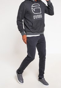 Felpa con cappuccio grigio scuro con un logo bianco "RAW", abbinata a jeans scuri e scarpe da ginnastica grigie, con un design a profilo basso e dettagli di cucitura a contrasto.