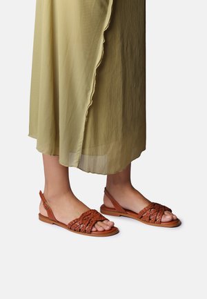 Naiste jalad, kes kannavad pruune punutud nahast lamejalanõusid ning oliivirohelist läbipaistvat seelikut lainelise äärega, valge tausta ees.