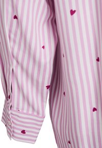 Chemise à rayures roses avec de petits motifs de cœurs rouges, fabriquée dans un tissu lisse, avec un détail de bouton sur le poignet et une fente sur le côté pour un ajustement décontracté.
