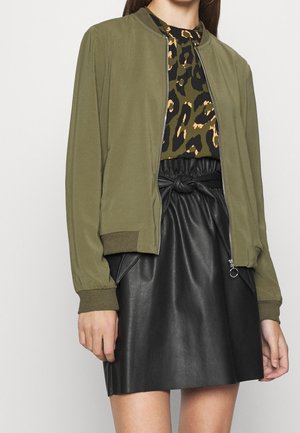 Femme portant une veste bomber olive sur un haut à imprimé léopard et une jupe en faux cuir noire avec un détail noué à la taille, posant devant un fond uni.