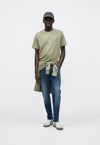 Oliwkowa zielona koszulka z logo, jasno szara bluza ze splotem, niebieskie dżinsy oraz białe trampki z czarnymi paskami. Czarna czapka z wzorem.