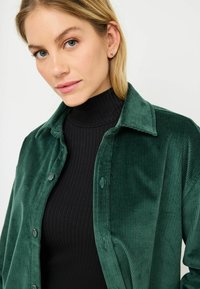 Groene corduroyjas met een knoopsluiting aan de voorkant en een gespreide kraag, gedragen over een nauwsluitende zwarte geribbelde coltrui. Gladde textuur, verticale strepen zichtbaar.