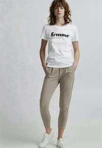 Camiseta branca de mangas curtas com o texto "femme" em preto, combinada com calças tapered bege e ténis brancos, em pé num fundo neutro.
