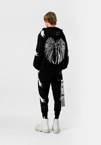 Persona con sudadera con capucha y pantalones negros con diseños de plumas blancas y botas blancas, de pie de espaldas contra un fondo liso.