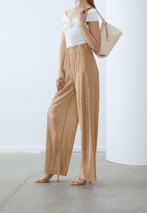 Pantalones anchos plisados beige, tela ligera, combinados con una blusa blanca sin hombros y sandalias de tacón alto con tiras. Bolso beige sencillo.