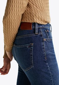 Modré denimové džíny s koženým nášivkem Tommy Hilfiger a lehkým prošíváním, spárované s texturovaným béžovým svetrem. Pohled z boku.