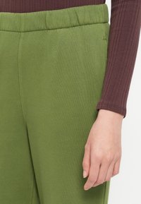 Pantalones de chándal verdes con cinturilla elástica, textura suave y bolsillos laterales. El conjunto se complementa con una blusa de color marrón oscuro con canalé.