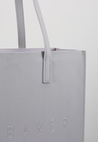 Sac fourre-tout texturé gris clair avec deux poignées et le mot « BAKER » embossé sur le panneau avant, sur un fond gris uni.