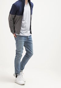 Námořnická a šedá mikina na zip, světle šedé tričko s véčkovým výstřihem, světle modré džíny a bílé sportovní boty s jemnou texturou a moderním střihem.