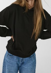 Sweat à capuche noir à coupe décontractée, avec des poignets et un ourlet côtelés. Associé à un jean en denim bleu clair. Texture lisse.