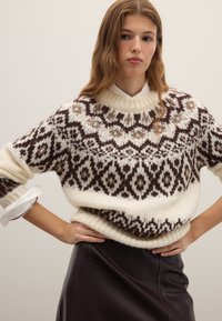 Gebreide trui in crème met een bruin en beige geometrisch patroon, voorzien van een geribde halslijn en manchetten, gecombineerd met een zwarte leren rok.