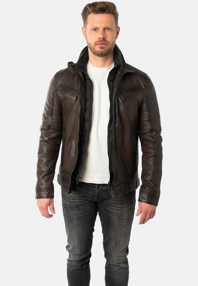 Chaqueta de cuero marrón con cuello alto, cremallera frontal y dos bolsillos en el pecho. Acabado texturado y forro interior acolchado. Llevada sobre una camisa blanca.