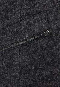 Strukturiertes schwarzes Gewebe mit hellgrauen Sprenkeln; verfügt über einen Reißverschluss und ein dezentes Steppmuster. Strapazierfähiges Material mit einer flauschigen Oberfläche.