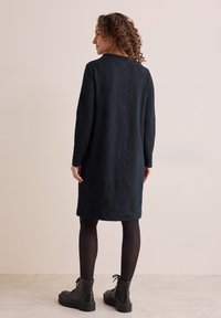 Robe noire texturée avec des manches longues et un col rond. Le tissu présente un motif ondulé en relief et arrive mi-cuisse. Bottines noires.