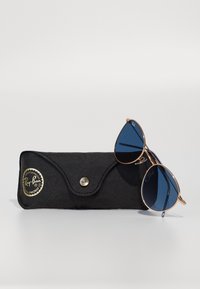Ochelari de soare Ray-Ban cu lentile albastre, ramă din metal auriu, odihnindu-se pe un toc din piele neagră cu închidere tip snap și detaliu cu logo.