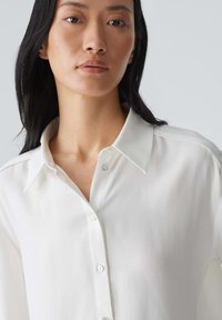 Chemise blanche à boutons avec un col classique, fabriquée en tissu lisse et léger. Présente des lignes épurées et un éclat subtil.