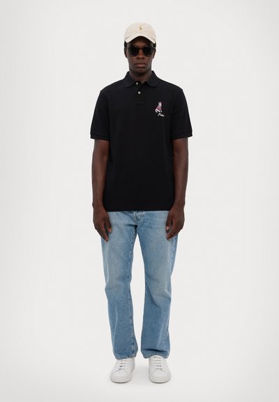 Cămașă polo neagră cu design brodat, blugi albaștri deschis, adidași albi și șapcă bej; accesorizată cu ochelari de soare negri.