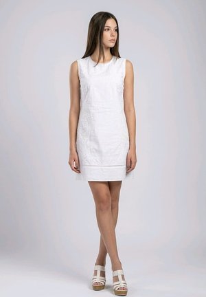 Mujer joven de pie con un vestido blanco bordado sin mangas y sandalias blancas de plataforma, mirando hacia su izquierda con un fondo liso.