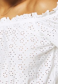 Blusa blanca sin hombros con tejido de ojales que presenta pequeños agujeros, escote con volantes y textura suave.