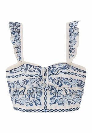 Femme portant un ensemble crème deux pièces avec des motifs floraux bleus, tenant un sac fourre-tout tressé avec des détails en cuir, debout devant un fond uni.