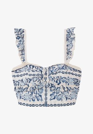 Mujer con conjunto crema de dos piezas con patrones florales azules, sosteniendo bolso tote tejido con detalles en cuero, de pie frente a fondo liso.