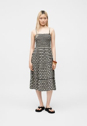 Jeune femme aux cheveux blonds et raides portant une robe midi noire à motif floral blanc, des sandales compensées noires et des bracelets en bois.