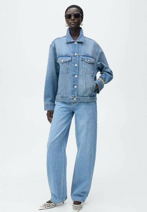Hellblaue Jeansjacke mit Kragen und vorderen Taschen, kombiniert mit dazu passenden weit geschnittenen Jeans. Das Model trägt schwarze Sonnenbrillen und gemusterte Schuhe.