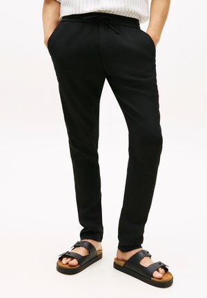 Osoba ubrana w czarne spodnie ze sznurkiem i czarne sandały na klamrę, stojąca z rękami w kieszeniach na jednolitym jasnym tle.