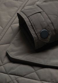Dark gray quilted jacket with a smooth texture, featuring a folded sleeve with a button accent and diagonal stitched pattern.