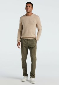 Beige gebreide trui met geribde manchetten en kraag, gecombineerd met olijfgroene cargobroek met zijzakken. Witte sneakers maken de outfit compleet.