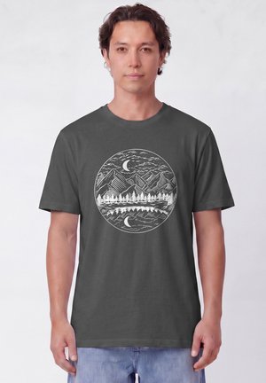 Mann trägt dunkelgraues T-Shirt mit weißem kreisförmigem Berg-, Baum- und Mondsichel-Design, das sich im Wasser spiegelt.