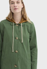 Veste légère verte avec une capuche marron, dotée d'une fermeture à boutons, d'un cordon de serrage et de poches latérales. La texture est lisse et semi-mate.